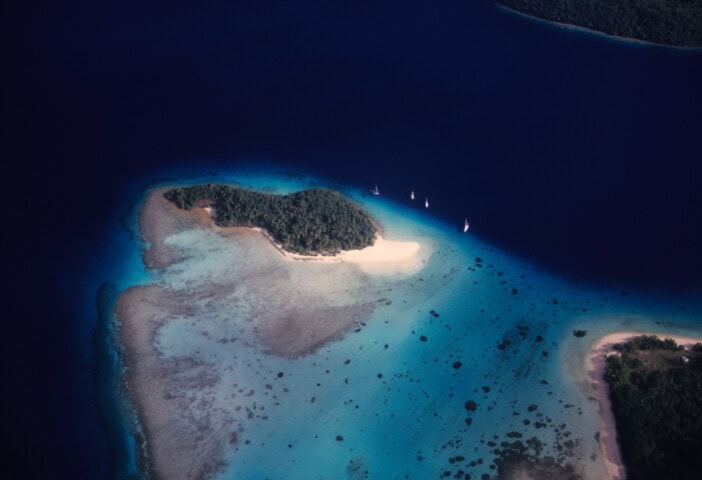 Aerial view of a small, forested island in the South Pacific surrounded by varying blue shades of water, with several boats anchored nearby.