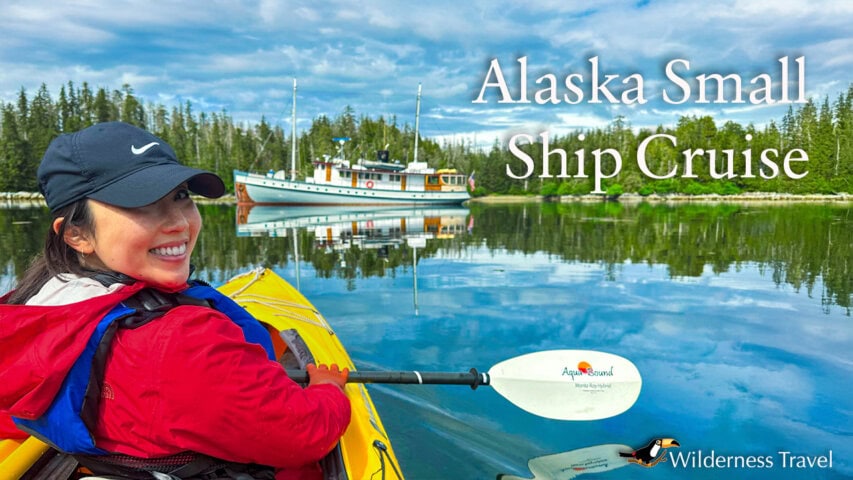 A woman in a red jacket kayaks on calm water near a small cruise ship, surrounded by forest, with "Alaska Small Ship Cruise" text and a Wilderness Travel logo.