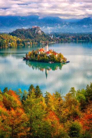 An island with a white church and red-roofed buildings sits in the middle of a serene lake surrounded by autumn foliage and distant mountains under a cloudy sky, making Slovenia an ideal travel destination for those seeking picturesque landscapes.