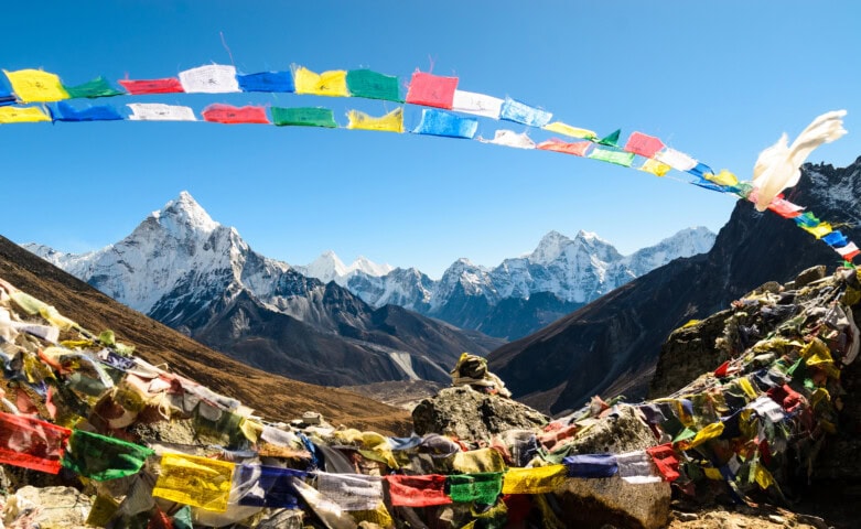 Colorful prayer flags flutter in the foreground, with a backdrop of Nepal's jagged snow-capped mountains under a clear blue sky.