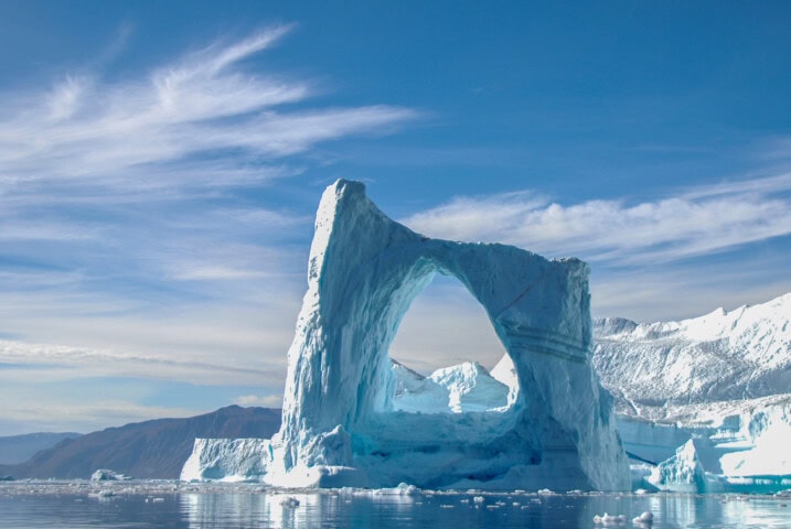 A large iceberg with a natural arch formation stands in the water against a backdrop of mountains and a partly cloudy sky, showcasing the breathtaking beauty of Greenland's landscape—a must-see for any travel enthusiast.