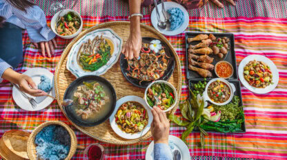 Several people sit around a woven mat, sharing a colorful spread of Thai dishes—a culinary adventure featuring rice, curries, grilled meats, vegetables, and dipping sauces.