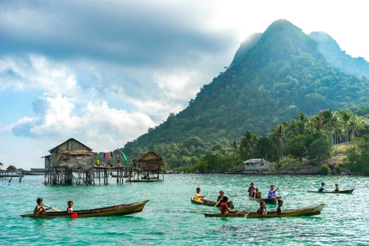 People in small wooden boats navigating turquoise waters, with stilt houses lining the shores and a lush, forested mountain in the background, capturing the picturesque essence of Malaysia.