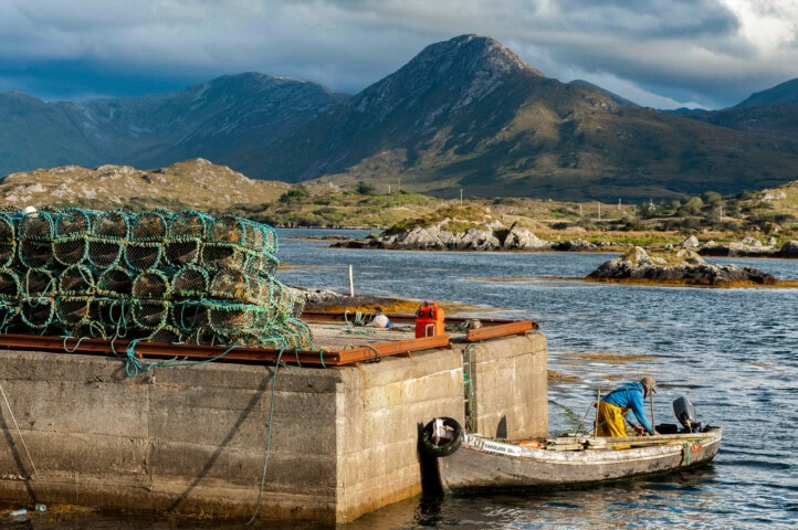 A person in a small fishing boat docks near a concrete pier stacked with lobster pots, with the rugged mountains and calm waters of Ireland providing a breathtaking backdrop.