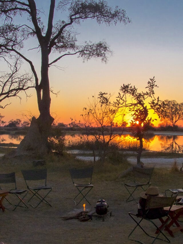 A serene campsite at sunset with empty folding chairs arranged in a circle around a small campfire, near a body of water. Silhouettes of trees and a clear sky enhance the peaceful setting, embodying the tranquil beauty of Botswana's landscape.