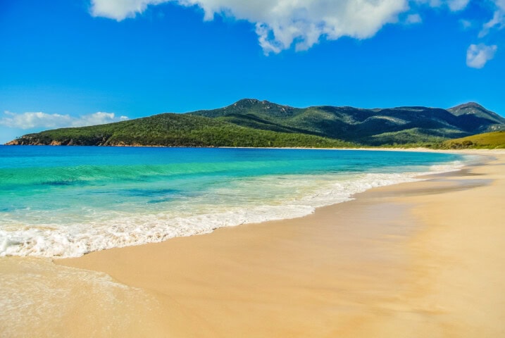 A serene Tasmanian beach with clear turquoise water and gentle waves, a sandy shore, and a backdrop of green mountains under a blue sky with scattered clouds—a must-visit spot for travel enthusiasts.