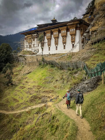 Two people walk along a narrow path towards a traditional hilltop building under renovation in Bhutan. The building is surrounded by scaffolding and wooden fences, with mountainous scenery in the background, capturing the essence of travel and tourism in this picturesque region.