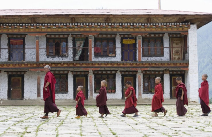 A group of monks, including young boys, walk in a line wearing red robes in front of a traditional two-story building with wooden windows and pillars—a serene scene that captures the essence of travel in Bhutan.
