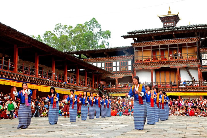 A group of women in traditional attire perform a synchronized dance in front of an audience at a picturesque Bhutanese monastery courtyard, featuring intricate wooden architecture and tiered balconies, making it a captivating experience for travel enthusiasts and tourism admirers alike.