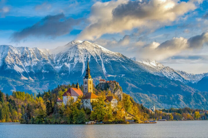 A small island with a church in the middle of a lake, surrounded by mountains with patches of snow under a partly cloudy sky, offers an idyllic setting perfect for travel enthusiasts. This picturesque spot in Slovenia is a hidden gem in the world of tourism.