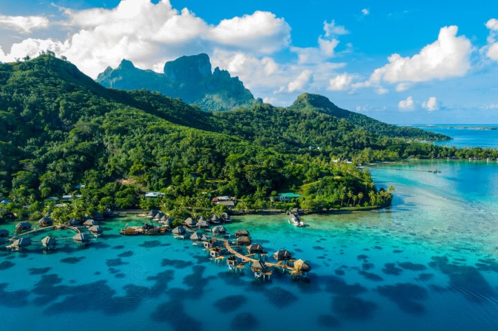 An overhead view of a tropical island in French Polynesia with lush greenery, overwater bungalows, clear blue waters, and distant mountains under a partially cloudy sky.