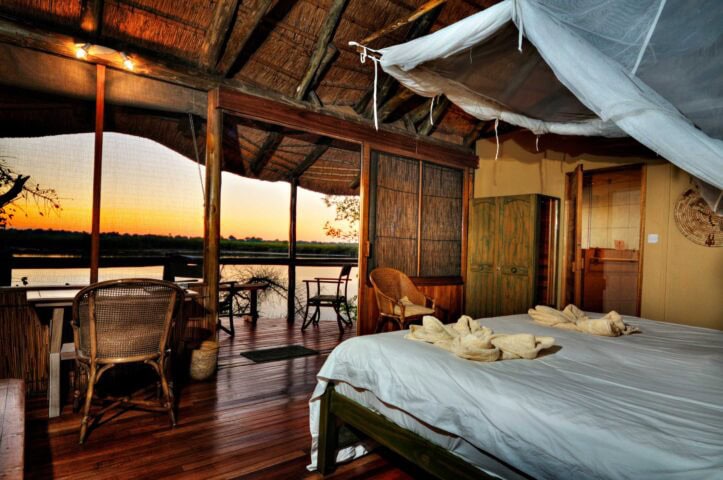 An open-air bedroom with a thatched roof, white sheets, and wooden furniture overlooks a scenic river and sunset through a large, screened window, offering the perfect setting for any traveler looking to relax after a day's hike.