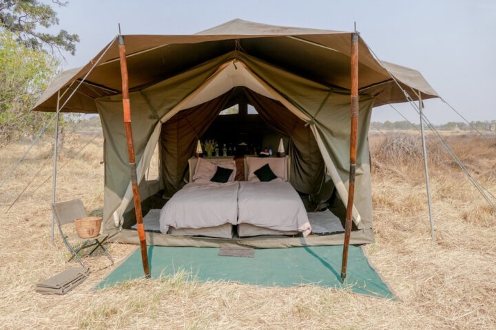A safari tent with two twin beds, a green mat at the entrance, and a small chair and basket outside, set in a dry grassy landscape, captures the best of Botswana.