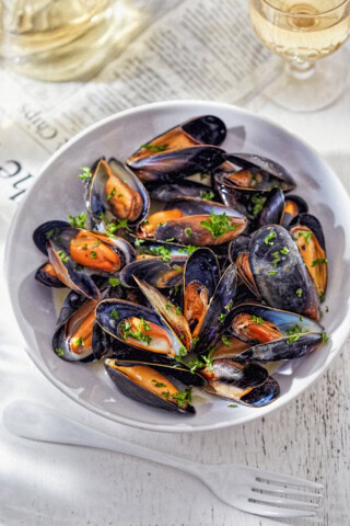 A white bowl with cooked mussels garnished with chopped parsley evokes the flavors of Ireland. A glass of white wine and a plastic fork are placed beside the bowl on a pristine white surface.