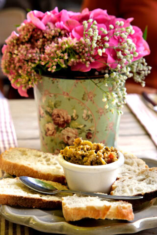 A small bowl of mixed olives sits on a plate with slices of bread, accompanied by a spoon. A metal floral vase with pink flowers is in the background, evoking the charm of a quaint café in France.