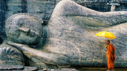A monk in orange robes holds a yellow umbrella while standing in front of a large stone reclining Buddha statue—a serene moment often witnessed on a Sri Lanka cultural tour.