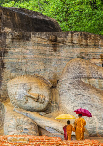 Two monks, one holding a yellow umbrella and the other a red one, stand in front of a large reclining Buddha statue carved into a rock wall in Sri Lanka, with lush greenery in the background.