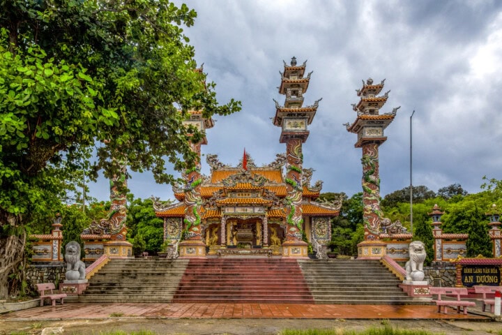 A vibrant, ornately decorated temple in Vietnam with multiple dragon-adorned columns and lion statues at the base steps. Flanked by large trees under a cloudy sky.