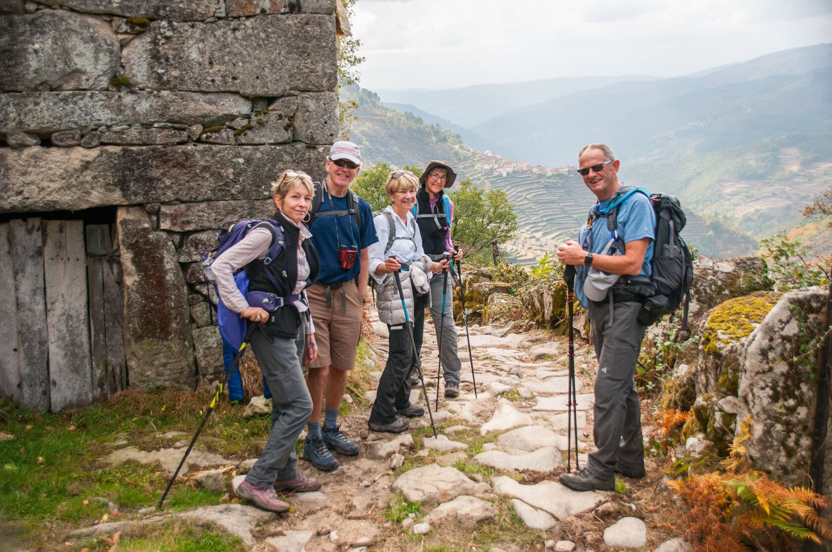 Five hikers with backpacks and trekking poles stand on a rocky path next to a stone building, overlooking terraced hills and a scenic valley—perfect for those seeking unique things to do in Portugal.