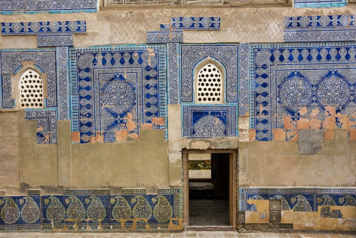 A wall adorned with intricate blue and white tilework featuring geometric and floral patterns, reminiscent of Uzbekistan's rich heritage, small arched windows with lattice screens, and a doorway revealing a view to another room.