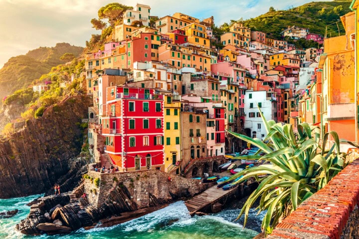 Colorful houses built on a rugged coastline in Italy, with boats docked at a small harbor. Green hills surround the village, and the ocean waves crash against the rocks below.