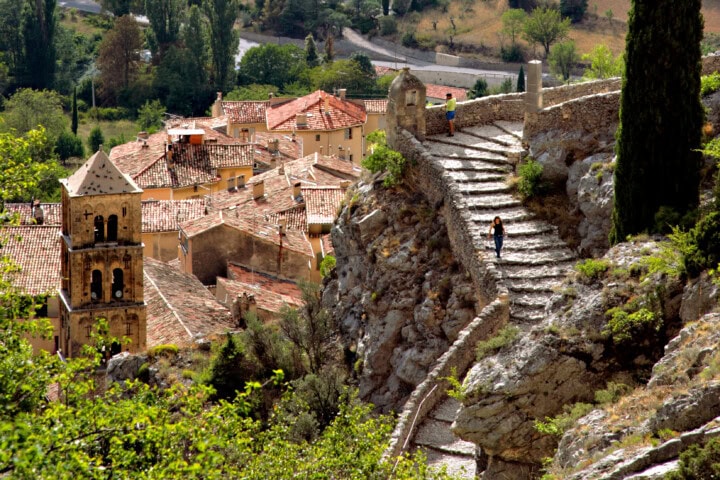 A person walks down a stone staircase on a hillside in France, overlooking a village with red-tiled roofs and a bell tower among trees.
