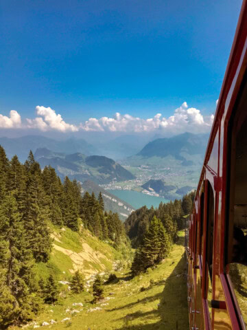 A red train travels through a picturesque mountainous landscape in Switzerland, with trees and a distant town visible under a bright blue sky with scattered clouds, offering an idyllic scene perfect for travel and tourism.