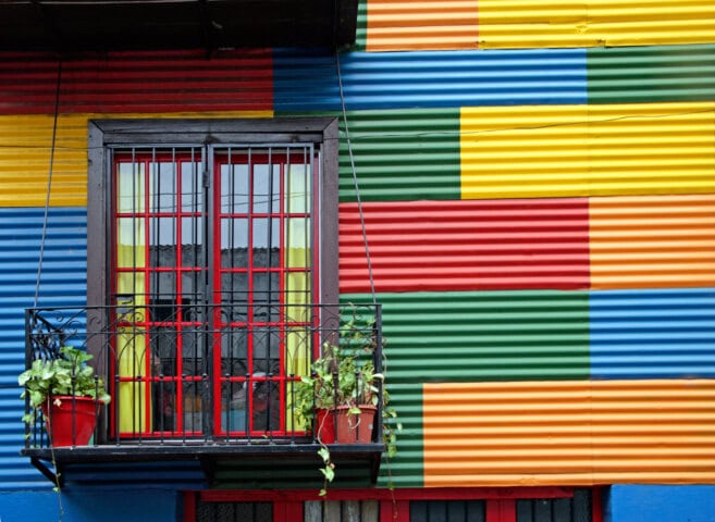 A window with metal bars and a small balcony adorned with potted plants is set against a building wall covered in multicolored corrugated metal panels, reminiscent of Argentina's vibrant architecture.