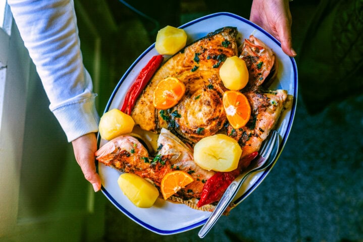 A person holds a platter of grilled fish fillets garnished with orange slices, cooked potatoes, and red peppers, reminiscent of a traditional Portuguese feast.