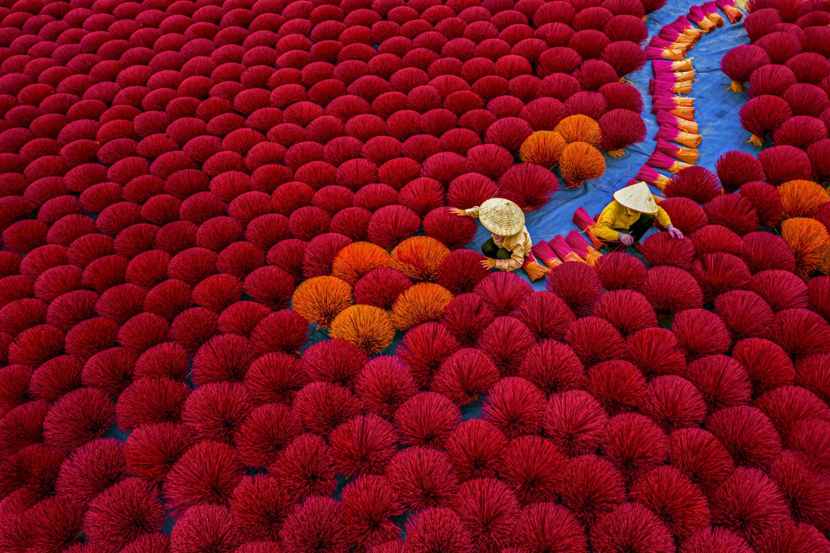 Two people wearing conical hats arrange bundles of red and orange incense sticks on a large outdoor drying area, creating a scene that feels like a vibrant page from a storybook about everyday heroes quietly preserving traditions at home.