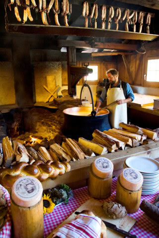 A person in an apron tends to a large pot in a rustic Swiss kitchen with hanging sausages and wood logs. Cheese wheels and bread are displayed in the foreground, evoking the charm of Switzerland for any travel enthusiast seeking authentic experiences.