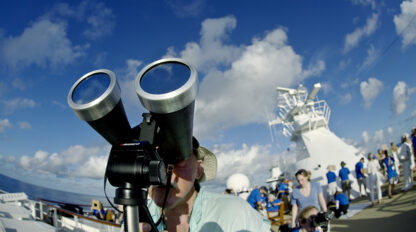 A person on a ship looks through large binoculars, marveling at the amazing sight in the universe, with other people and nautical equipment visible in the background under a blue sky with clouds.