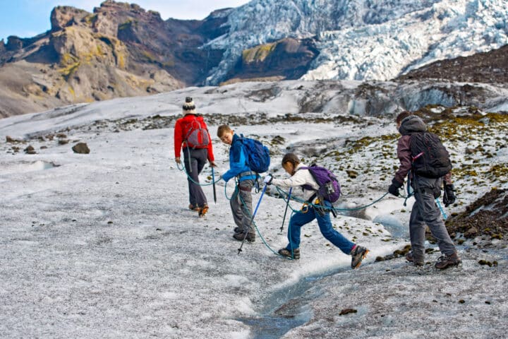 Four people wearing hiking gear and backpacks are roped together, trekking across an Argentinian glacier with rocky mountains in the background.