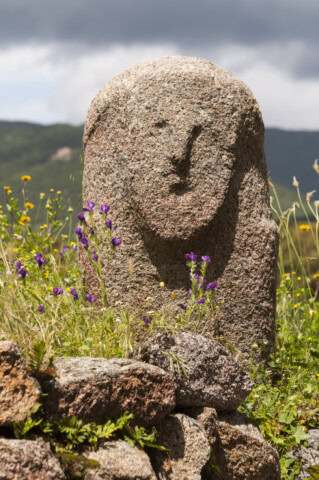 A weathered stone statue with a human-like face sits among rocks and wildflowers in an idyllic French outdoor setting, with majestic mountains in the background.