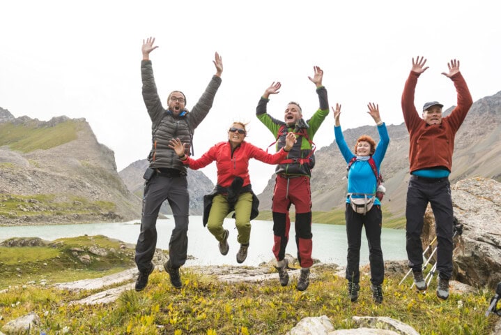 Five people are jumping in unison, dressed in outdoor gear with a stunning mountainous landscape and the serene lakes of Kyrgyzstan in the background.
