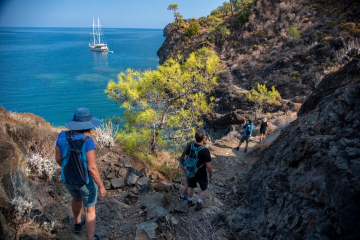 Four people hike down a rocky trail in Turkey towards a calm blue sea with a sailboat anchored near the shore.