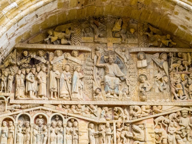 Detailed stone carvings on a French church portal depict religious scenes, including figures in various poses around a central figure on a throne, surrounded by angels, and inscriptions around the scenes.