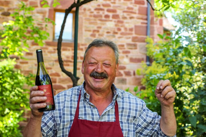 A man in a checkered shirt and red apron holds a bottle of wine while smiling with a stone building and lush greenery in the background, evoking the charm of French tourism.