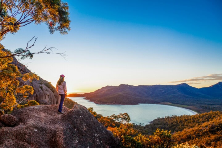 A person stands on a rocky ledge overlooking a scenic Australian landscape with a body of water, forest, and mountains under a clear blue sky at sunset.