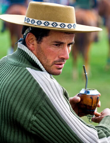 A man wearing a traditional hat and green sweater is holding a cup of mate in an outdoor setting, capturing the essence of Argentina's rich culture.