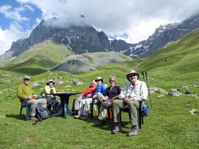 A group of six people in hiking gear sits around a table outdoors in a mountainous area, showcasing the stunning green fields and rocky peaks of Georgia, inviting an unforgettable travel experience.