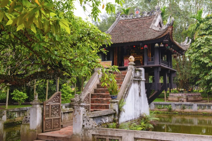 A historic Vietnamese pagoda with a red tiled roof, elevated on stone pillars above a pond, surrounded by lush greenery. Stone steps and a small gate lead up to this enchanting structure, embodying the serene beauty of Vietnam.