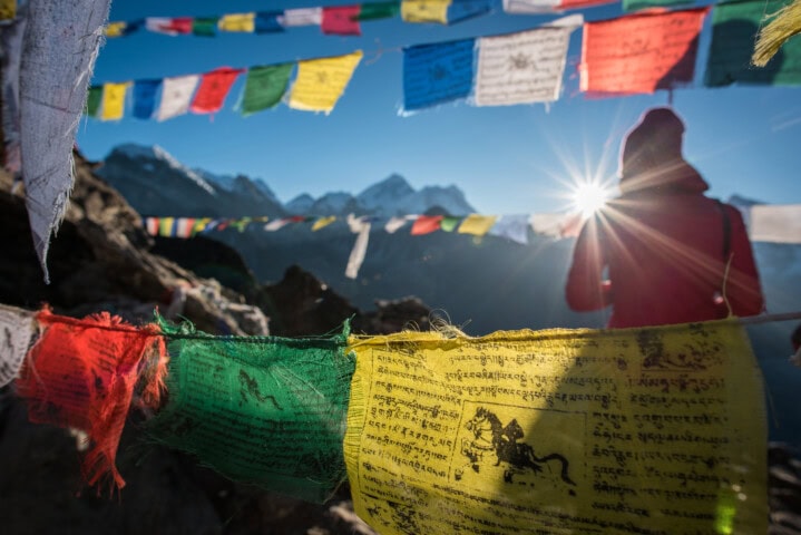 Colorful prayer flags flutter in the wind with snowy mountains in the background. A person in warm clothing stands silhouetted against the sun, capturing a moment of serene beauty often found in Nepal's Himalayas, a true gem for tourism seekers.