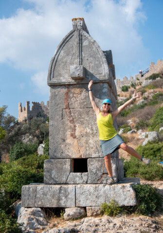 A person in a yellow top and blue cap poses energetically next to a large stone monument with a historical fort in Turkey visible in the background.