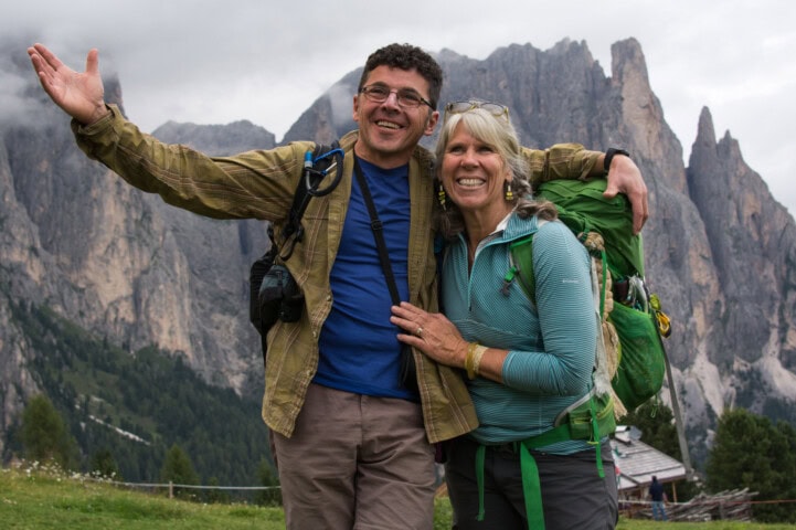 Two hikers with backpacks smile and pose with arms raised in front of a stunning mountainous landscape in Italy.