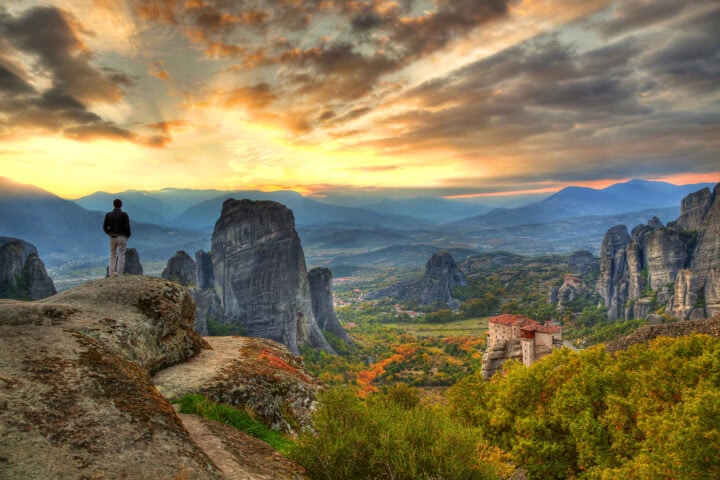 A person stands on a rocky edge overlooking a Greece valley with tall rock formations and a stunning sunset sky. A small building is visible among the trees.