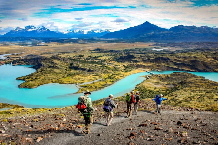 A group of hikers walks on a trail overlooking a landscape in Argentina, with turquoise lakes, rugged hills, and snow-capped mountains under a partly cloudy sky.