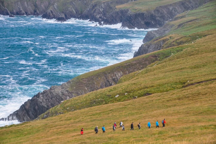 A group of people is hiking on a grassy hillside in Ireland, overlooking a rocky coastline and ocean waves.