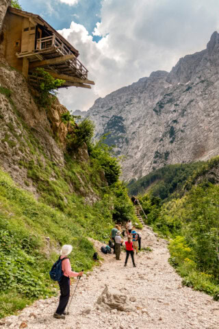 Hikers walk on a rocky mountain trail in Slovenia, with a wooden cabin perched on a cliff above them and a breathtaking mountainous landscape in the background, showcasing the allure of travel and tourism.