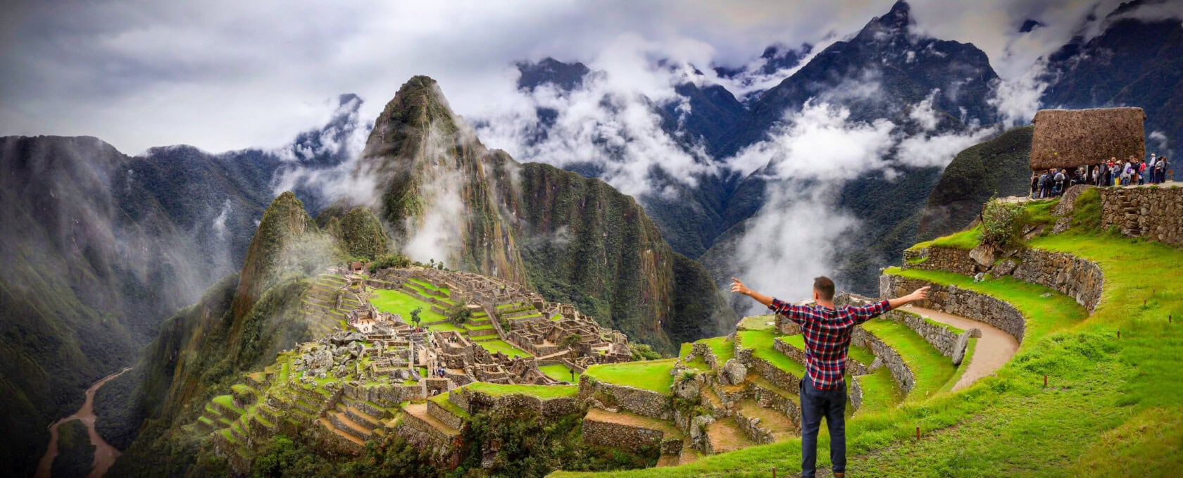 A person stands with arms outstretched overlooking the ancient Incan city of Machu Picchu, surrounded by green mountains and misty clouds.